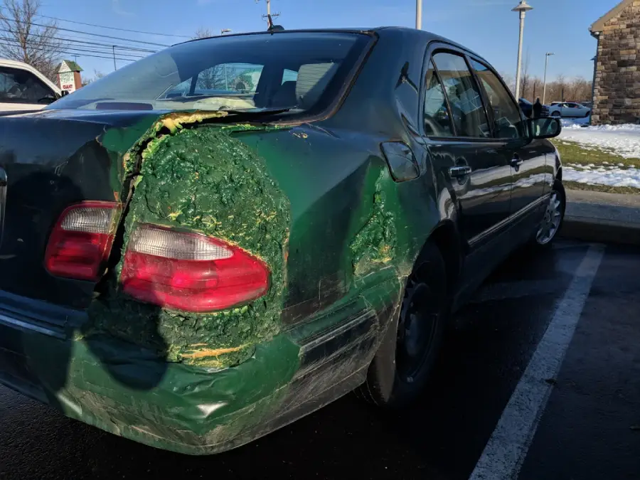 A green car with significant damage to the left rear side, patched roughly with green foam or filler. The repair is uneven, and the car is parked in a lot with some snow and buildings visible in the background.
