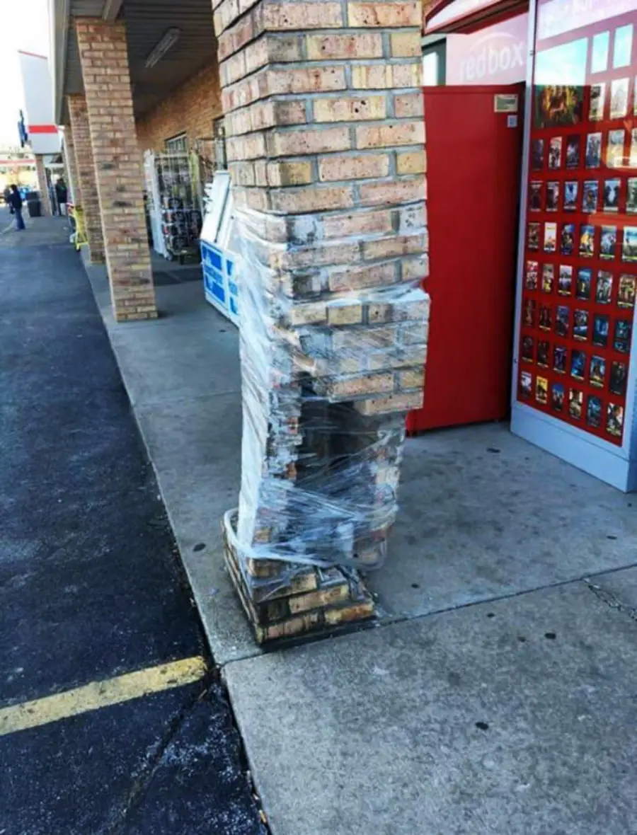 A damaged brick pillar wrapped in plastic wrap stands outside a store next to a red movie rental kiosk, showing crumbling bricks at its base.