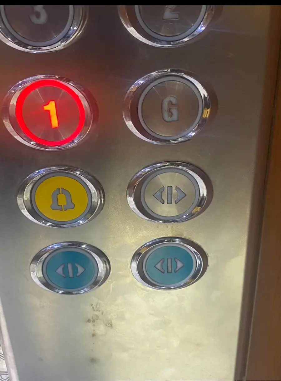 Close-up of elevator buttons: “1” is lit up in red, “G” is above it. Below are yellow, gray, and blue buttons for a bell, door close, and door open functions. Stainless steel panel.