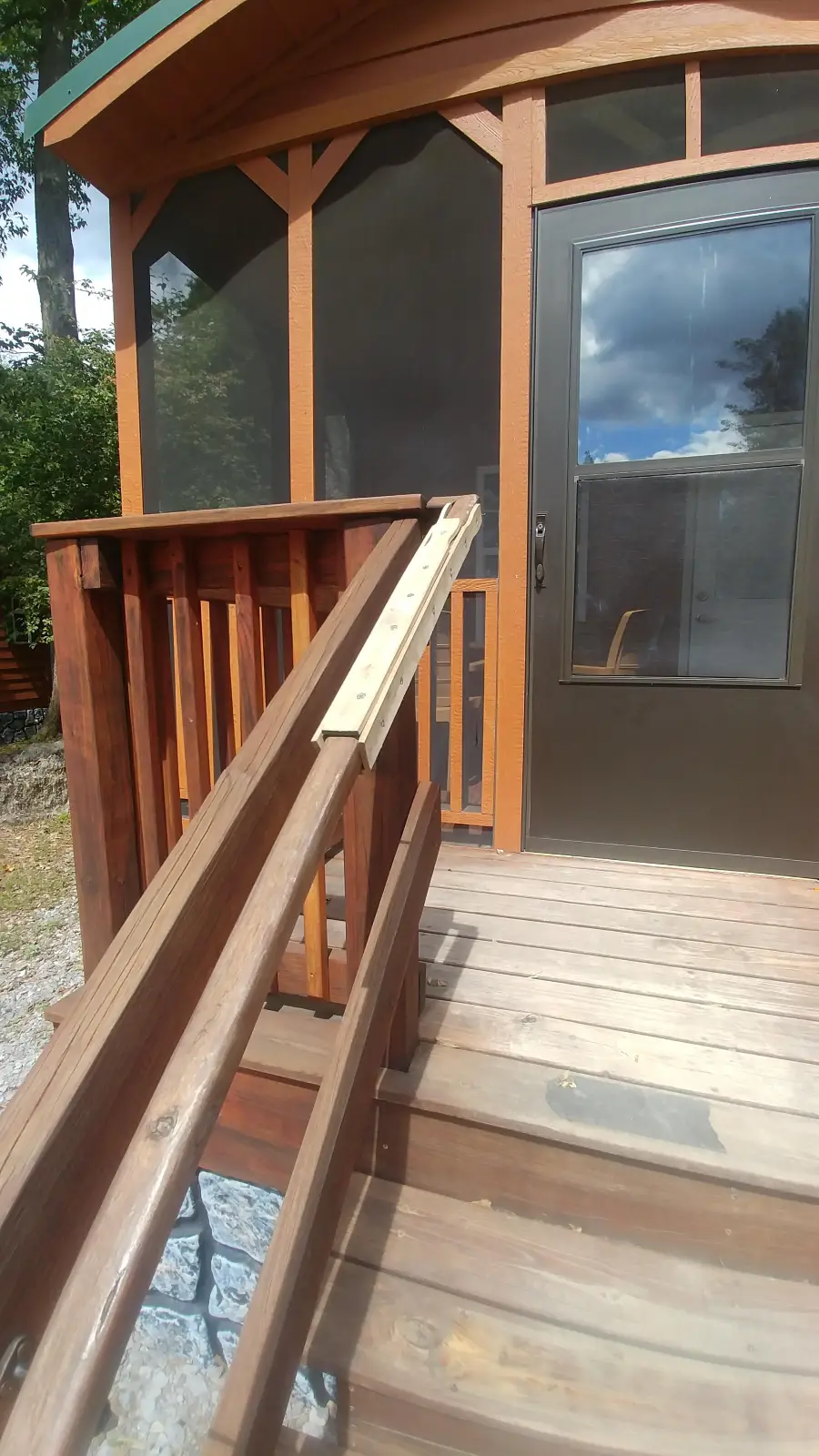 A wooden ramp is attached to a stair railing leading up to a screened porch with a glass door, surrounded by trees. The weather is sunny with some clouds.