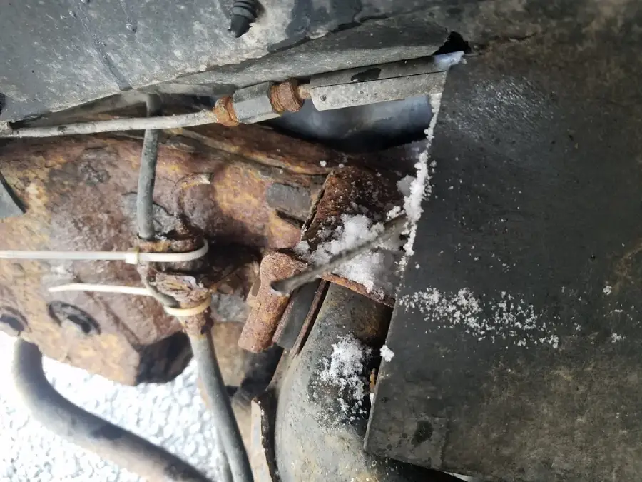 A close-up view of a rusted car suspension or brake component with some snow and ice buildup, metal rods, and zip ties visible.