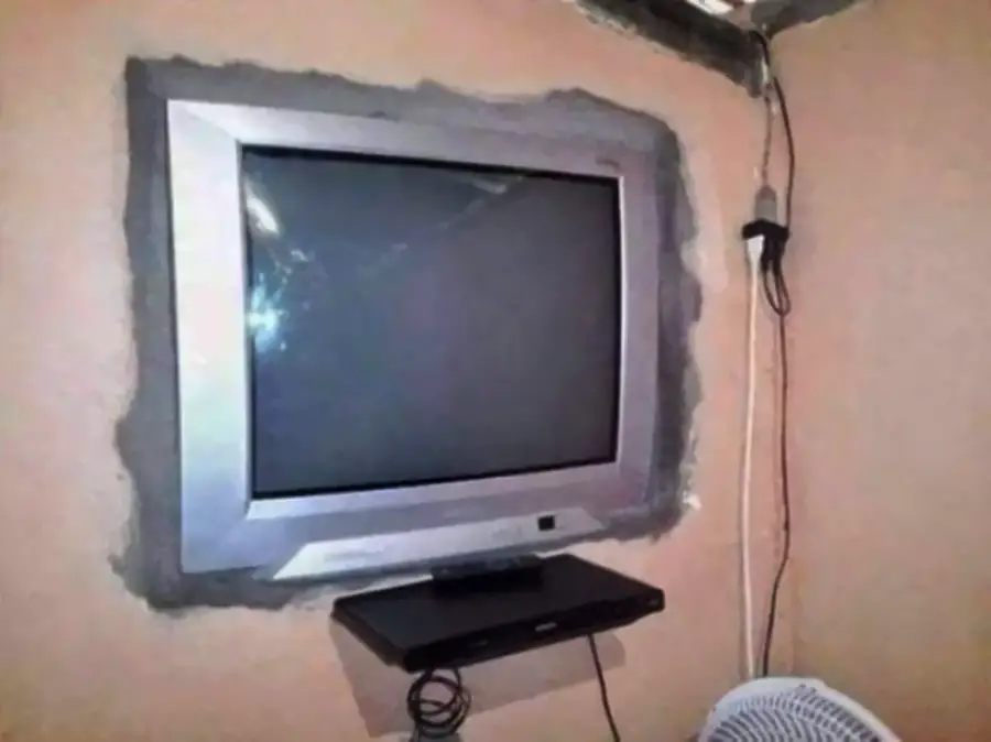A flat-screen TV is mounted inside a wall with rough cement edges. Below it, a black electronic device sits on a small shelf. Wires hang to the side, and part of a white fan is visible in the bottom right corner.