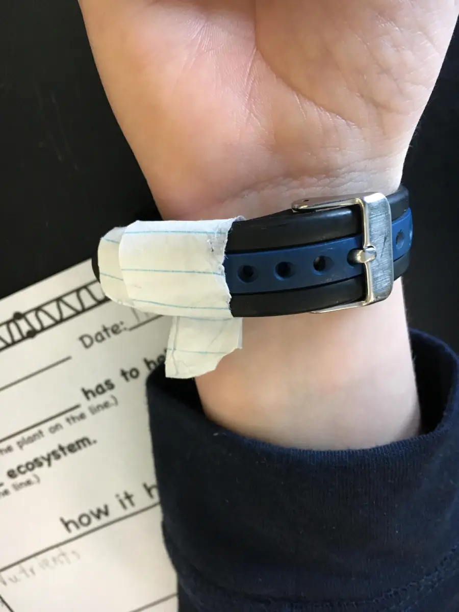 A hand wears a watch with a strap fastened by a piece of lined paper. Part of a worksheet with the words “how it h” is visible next to the wrist.