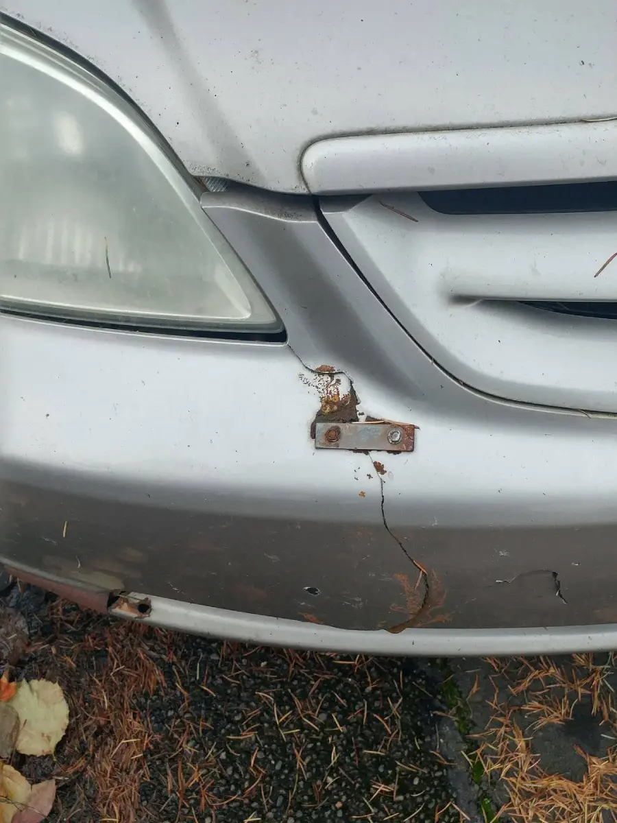 A close-up of a silver car’s front, showing a cracked bumper held together by a rusted metal bracket and screws. Pine needles and leaves are on the ground nearby.