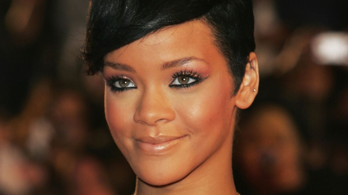A close-up portrait of Rihanna with short, dark hair, light brown eyes, and neutral makeup, smiling softly at the camera against a blurred dark background.