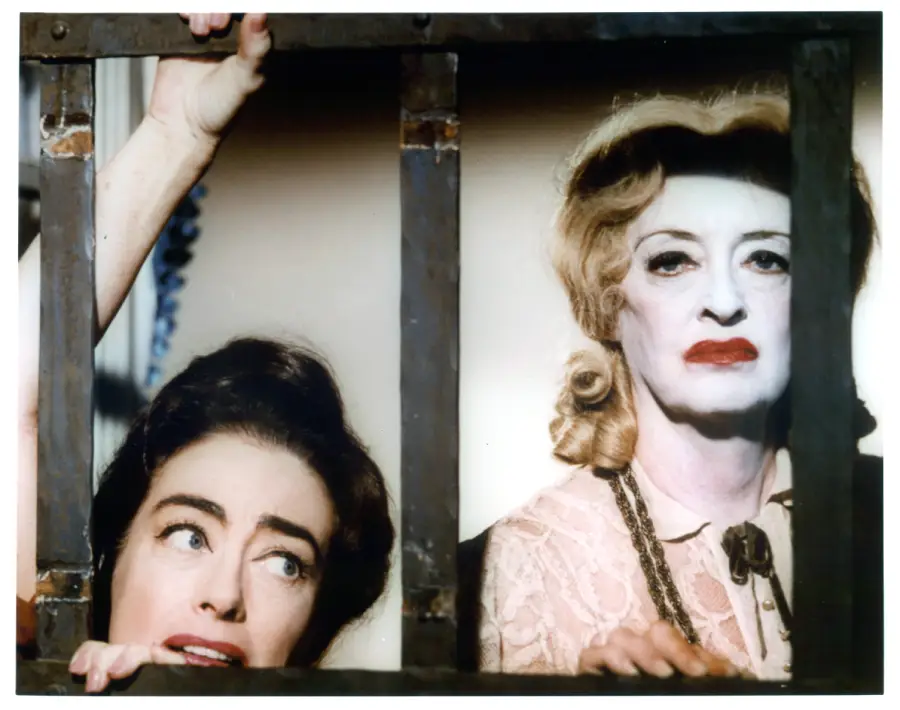 Two women framed by window bars; one looks up anxiously with dark hair, while the other, older and heavily made up with curly blonde hair, looks forward with a stern expression.