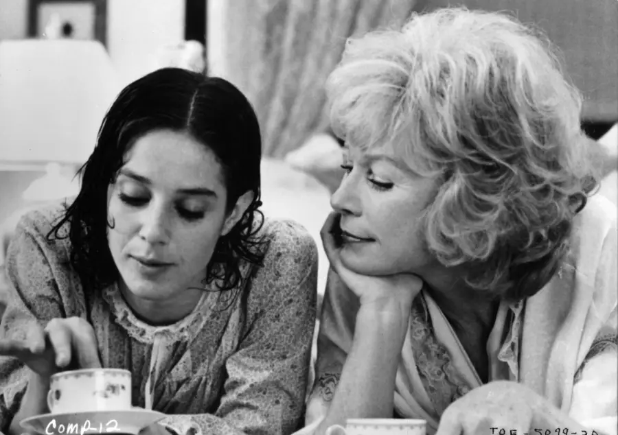 Two women sit close together at a table, sharing a quiet moment. One woman looks down at a teacup, while the other, with lighter hair, leans on her hand and gazes at her companion with a gentle expression.
