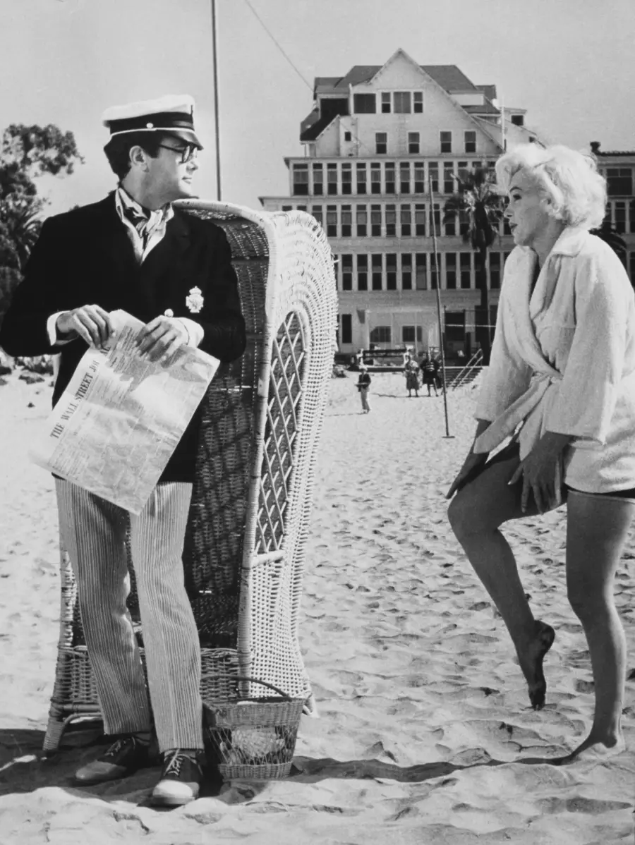 A man in a sailor hat and blazer holds a newspaper on a beach, standing beside a wicker chair, as a woman in a robe and swimsuit playfully lifts one leg near him. A large building stands in the background.