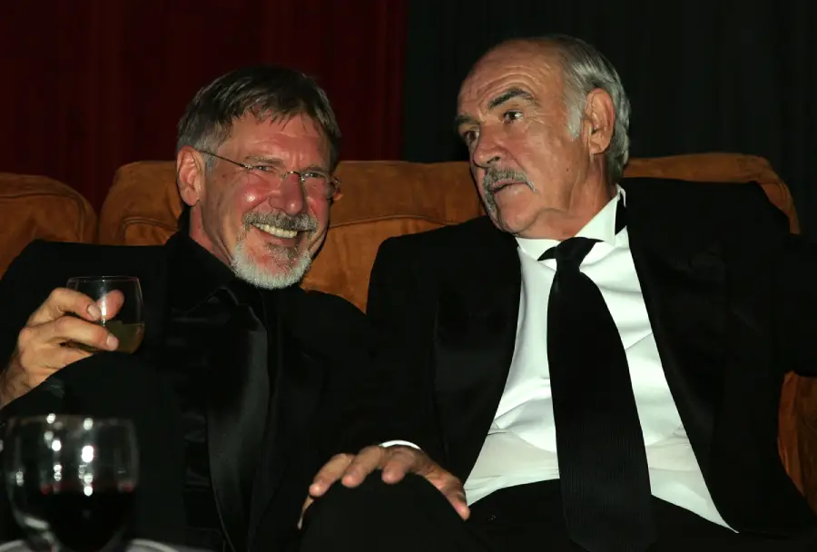 Two older men in black suits sit closely on a brown couch, smiling and talking. One holds a drink. The background is dark, and there’s a glass of red wine on a table in the foreground.