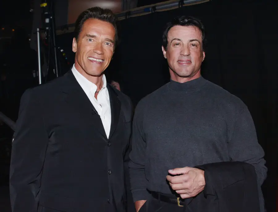 Two men stand together, smiling at the camera. One wears a black suit with a white shirt, and the other wears a dark sweater and holds a jacket. The background is dark and out of focus.