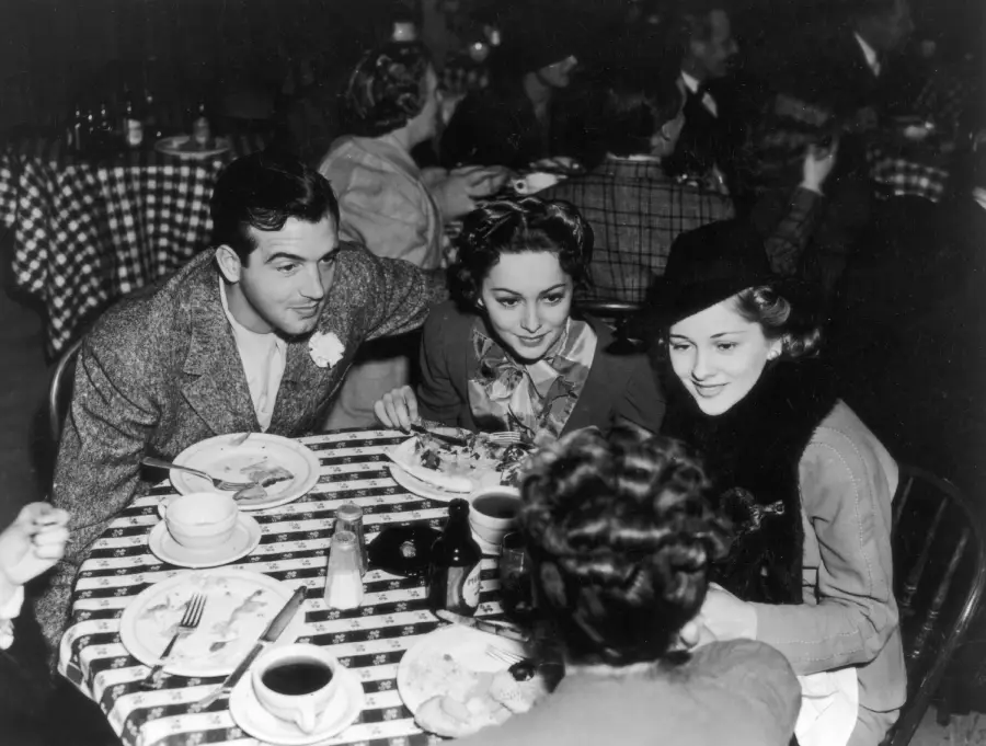 Four people sit around a table with checkered tablecloths, eating and talking in a busy restaurant. Plates, cups, and utensils are on the table. The photo is black and white, with a vintage, classic atmosphere.