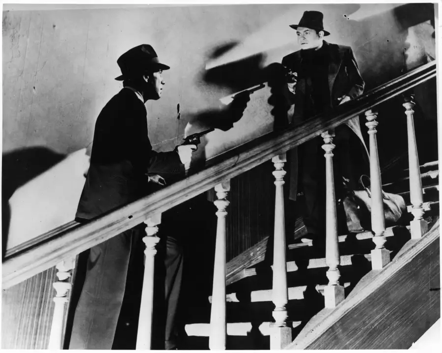 Two men in suits and hats point guns at each other on opposite sides of a staircase, with dramatic shadows cast on the wall behind them in a black-and-white scene.