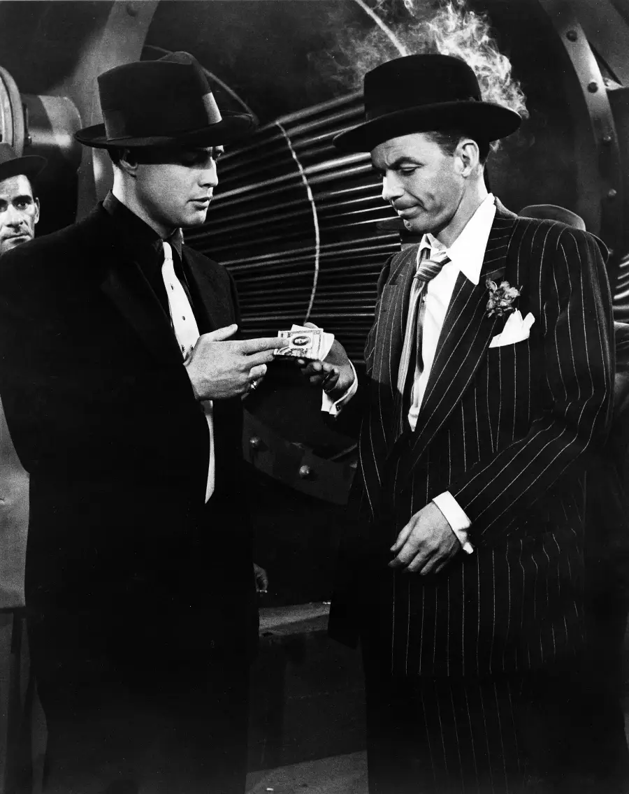 Two men in 1940s-style suits and hats stand together; one hands a small item to the other. Smoke rises from behind the man on the right. Industrial machinery is visible in the background.