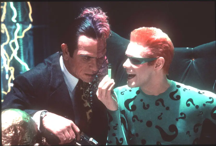 Two men in costumes: one with a split face and suit, the other with bright orange hair in a green, question mark-patterned outfit, holding a small clear vial and grinning. Neon lights glow in the background.