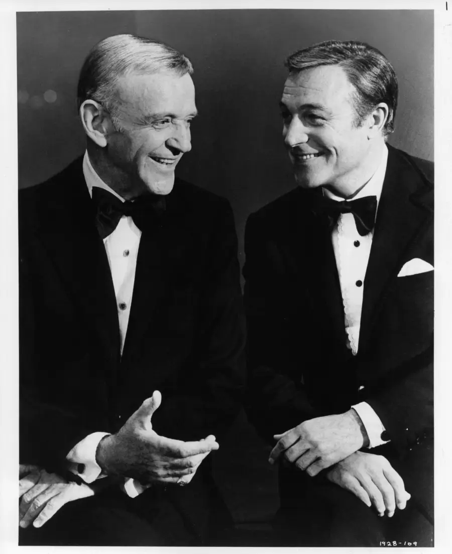 Two men in tuxedos sit side by side, smiling at each other against a plain dark background. Both have neatly styled hair and are engaged in friendly conversation.