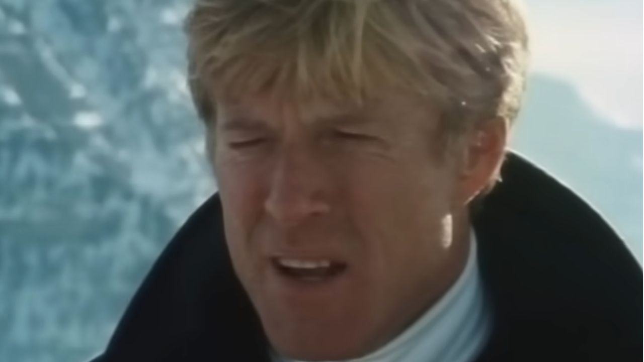 A man with blond hair wearing a dark coat and white turtleneck stands outside in a snowy, mountainous setting, squinting slightly as he looks ahead.