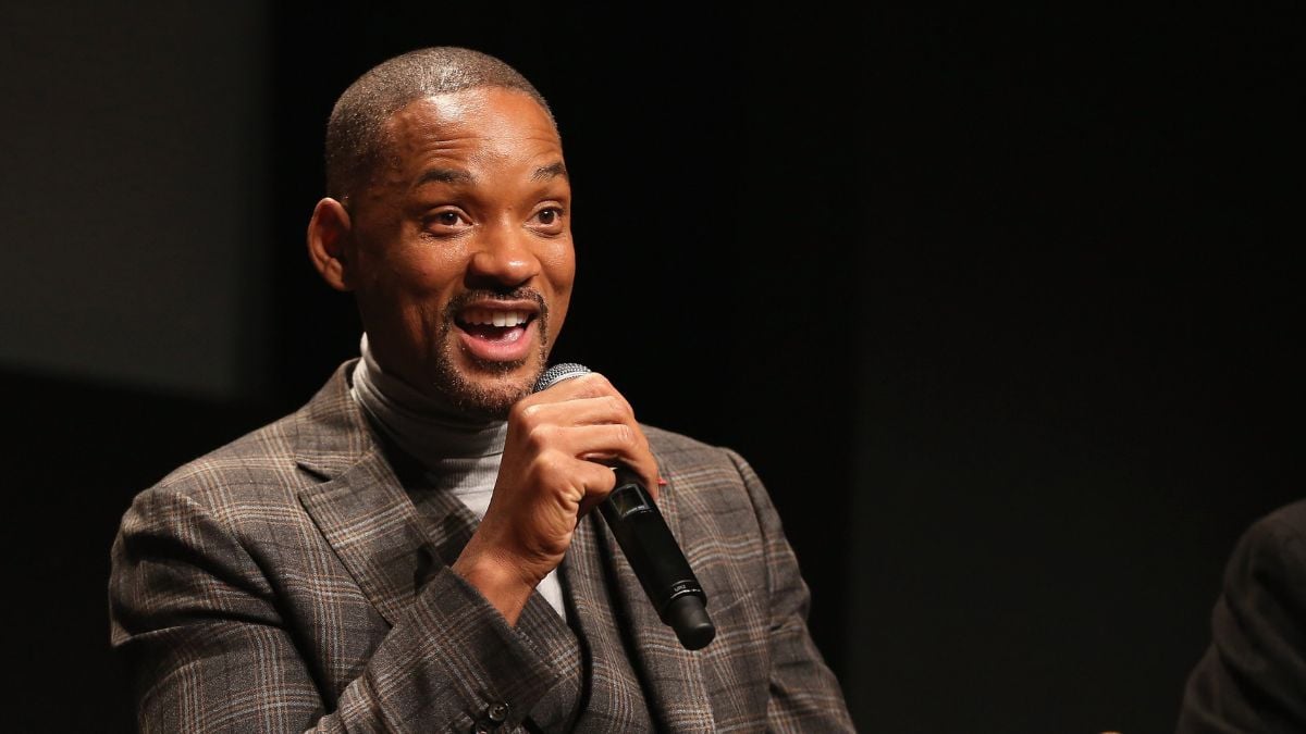 A man in a plaid suit speaks enthusiastically into a microphone while gesturing with his hand; he is sitting against a dark background.
