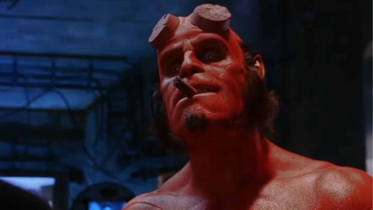 A red-skinned character with filed-down horns, sideburns, and a cigar in his mouth stands shirtless in a dimly lit, industrial setting.