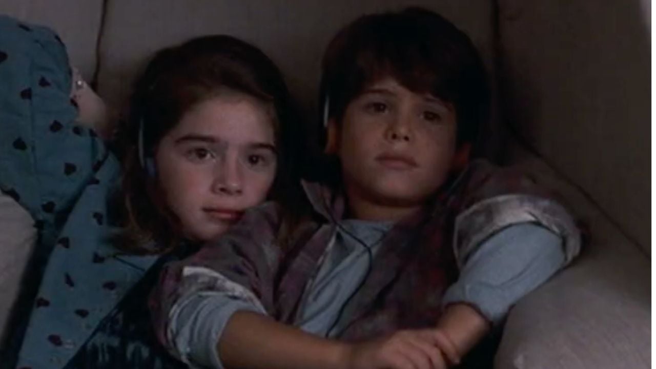 Two young children, a girl and a boy, are sitting close together on a couch, looking ahead with calm expressions. They both have brown hair and are wearing casual clothes.