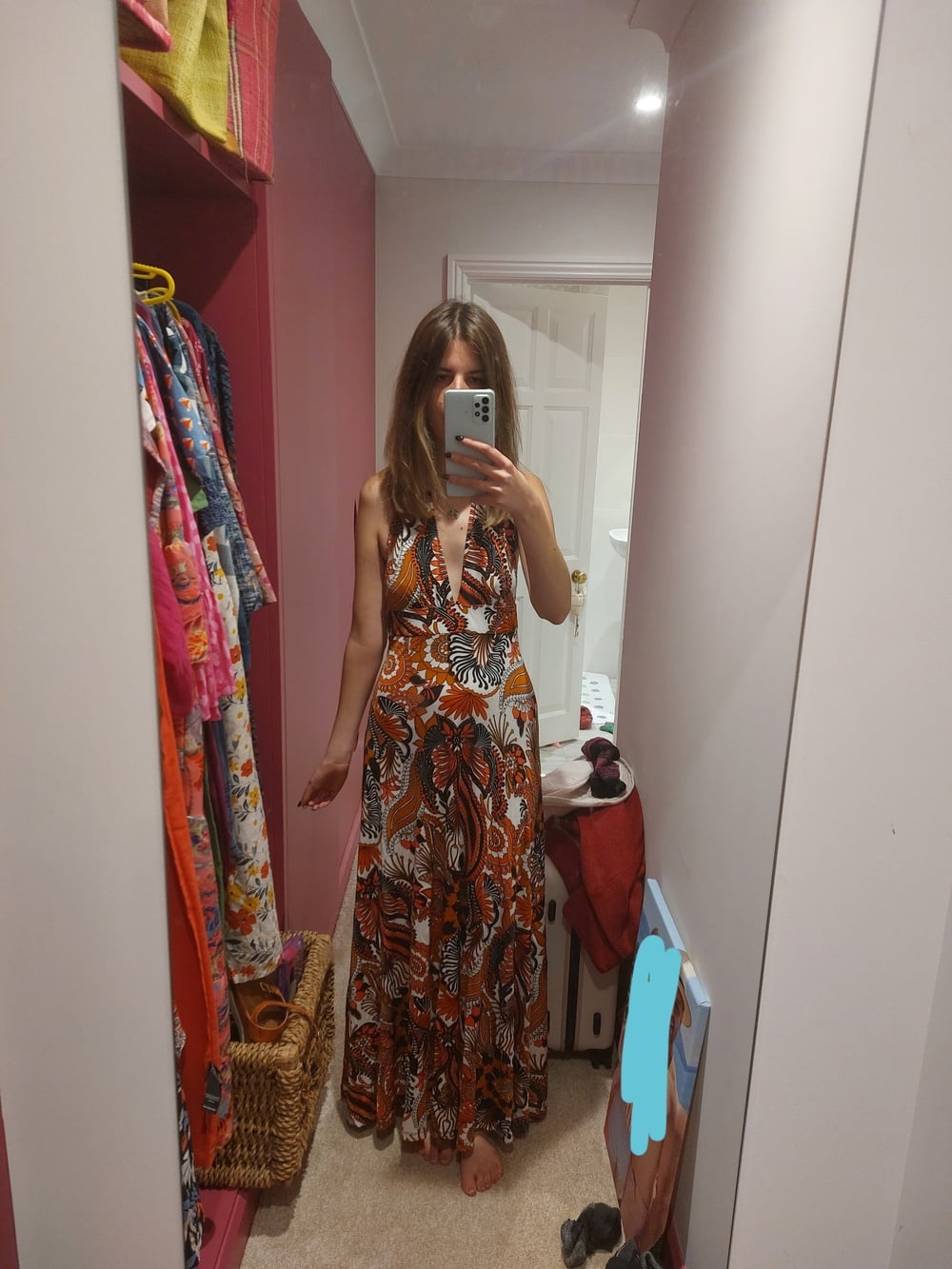 A woman stands barefoot in front of a mirror, wearing a long, colorful, patterned halter dress. The room has pink walls, a rack of clothes, and a basket on the floor. She is holding a phone to take a mirror selfie.