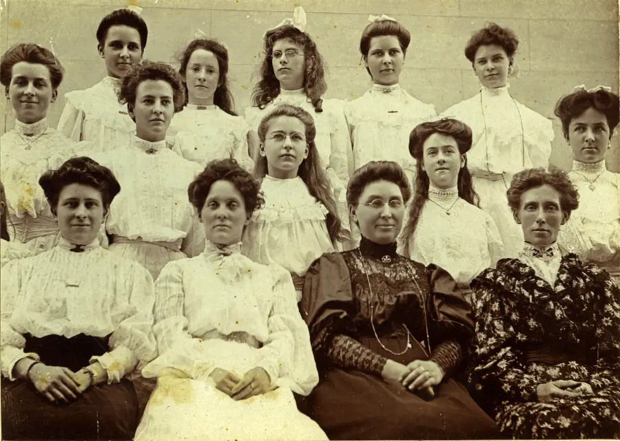 Fourteen women in formal, old-fashioned dresses pose for a group portrait; most are wearing high-necked blouses with lace details, and their hair is styled up or in waves. The photo appears sepia-toned and vintage.