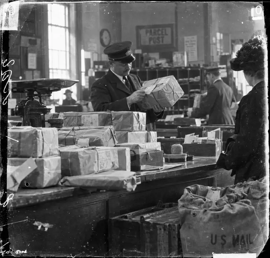 Black and white photo of a postal worker examining a package in a busy post office, surrounded by parcels, scales, and mail bags. Signs reading “PARCEL POST” and “U.S. MAIL” are visible in the background.