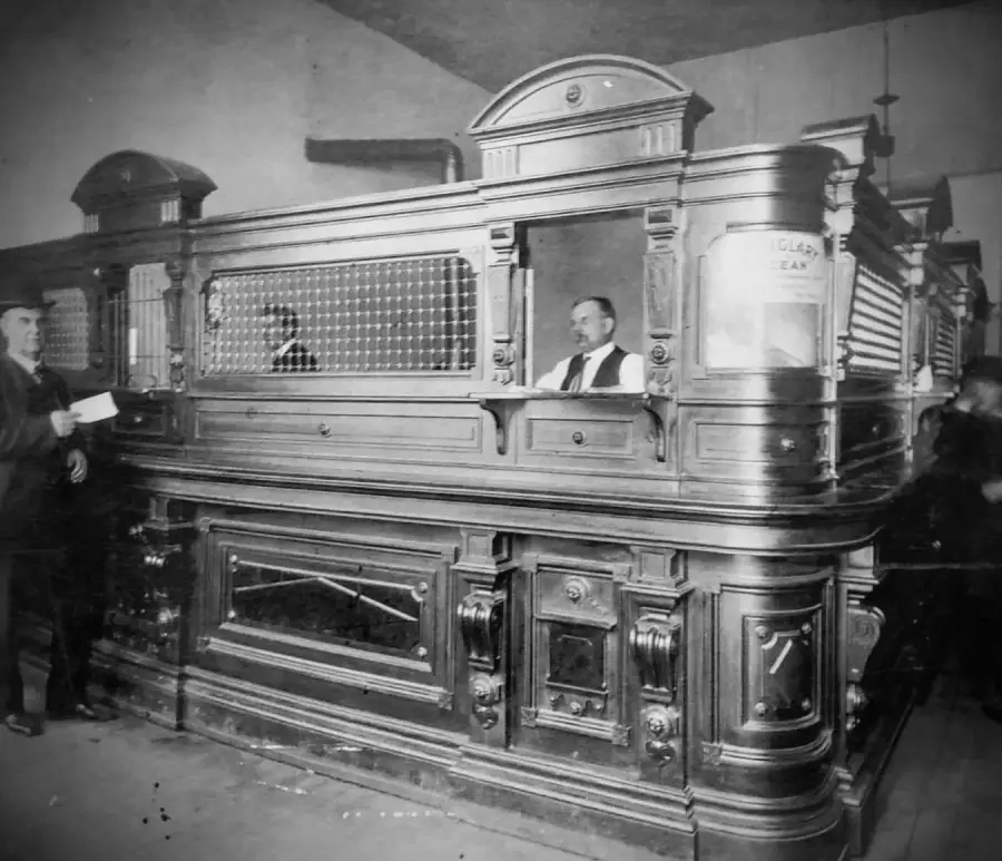 A black-and-white photo of an ornate, vintage bank teller counter with a teller inside and a customer handing over documents. The counter features decorative woodwork and glass panels.