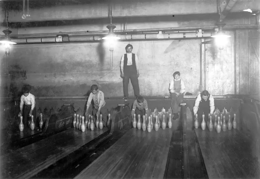Five boys manually reset bowling pins in a vintage bowling alley. One man stands behind, while the boys crouch or sit on the lanes, working under exposed pipes and hanging lights. The scene is black and white.