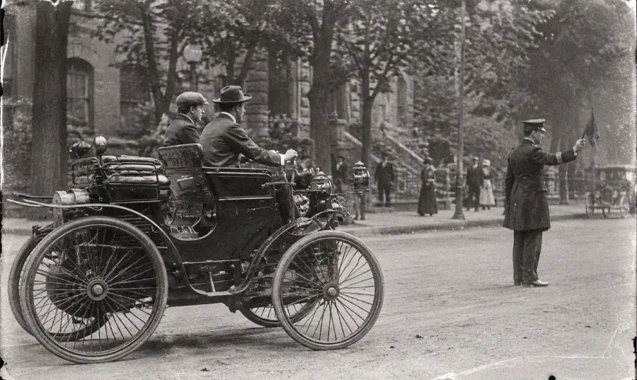 A vintage black-and-white photo shows an early automobile with two men inside, stopped as a uniformed traffic officer directs vehicles on a city street lined with trees and pedestrians.