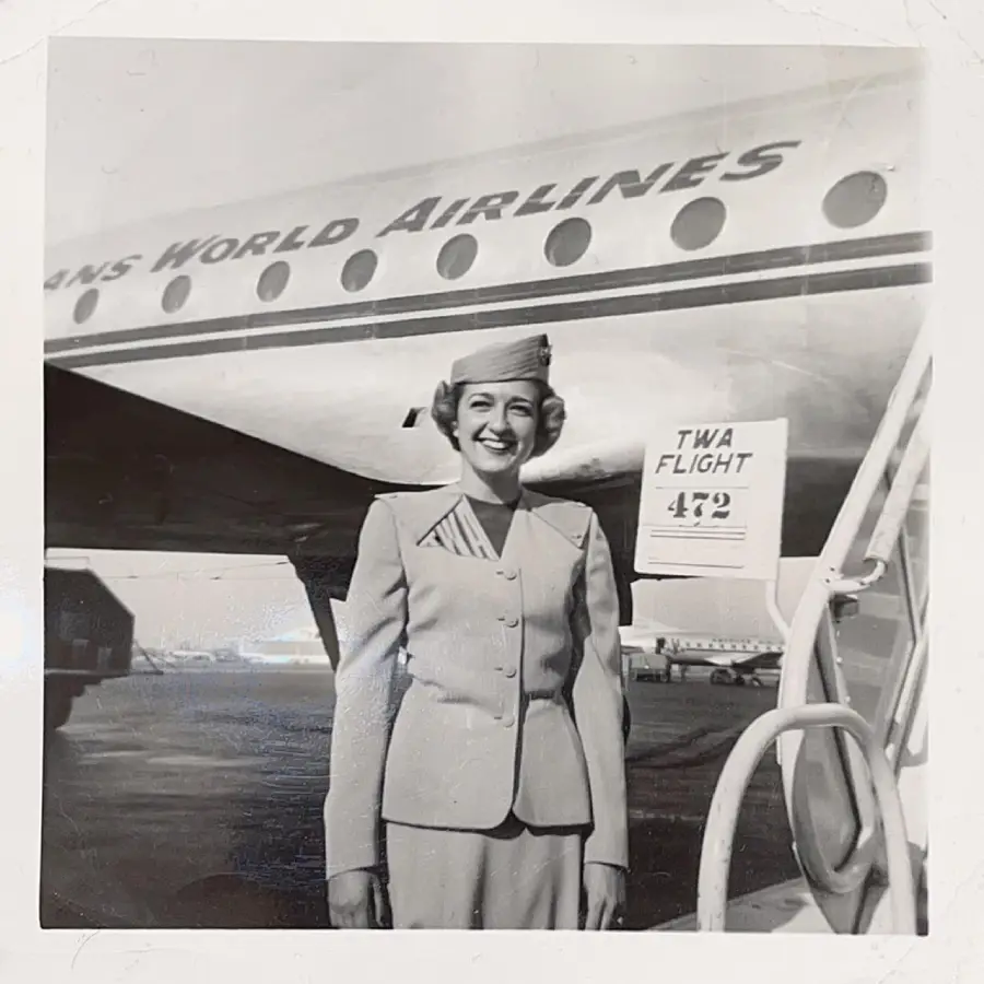 A smiling flight attendant in a vintage uniform stands in front of a Trans World Airlines airplane near a sign that reads “TWA Flight 482.” Steps lead up to the plane’s entrance. The image is black and white.