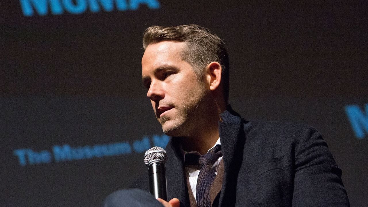 A man in a dark coat holds a microphone while sitting and speaking at an event. The background is dark with partially visible blue text.