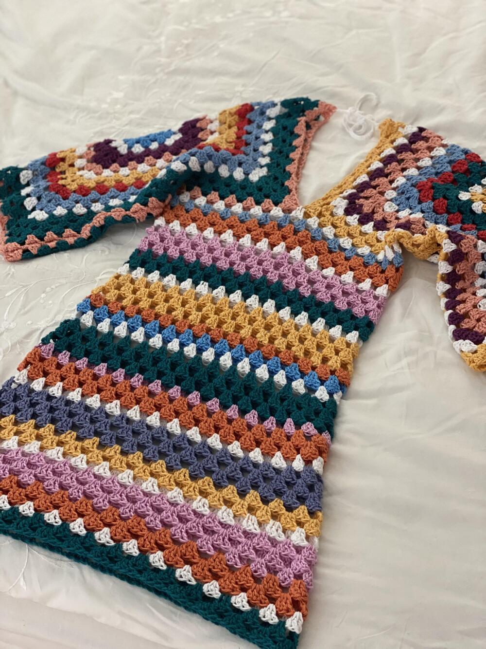 A colorful, handmade crochet dress with multicolored stripes and granny square-patterned sleeves, laid flat on a white surface.