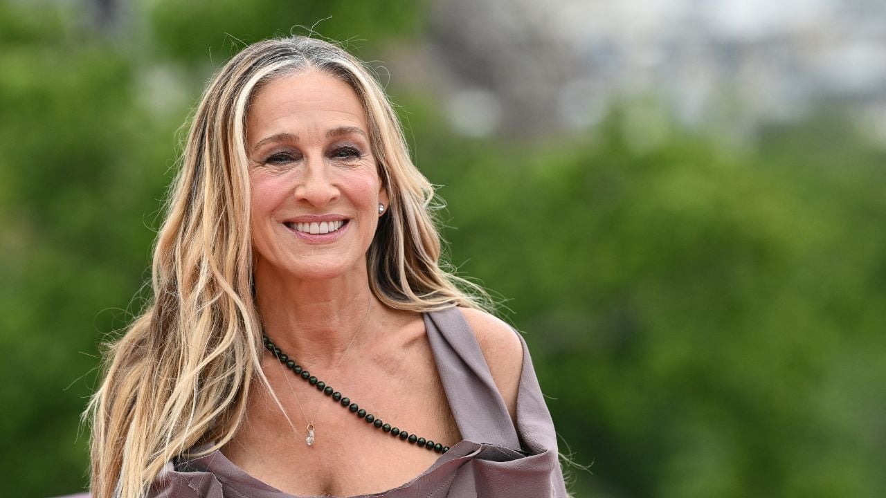 A smiling woman with long, wavy blonde hair stands outdoors, wearing an off-shoulder taupe dress and a black beaded necklace, with blurred greenery in the background.