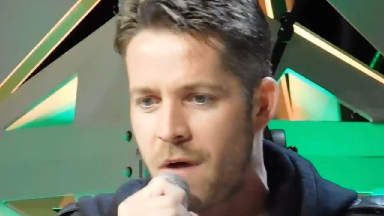 A man with short brown hair and blue eyes holds a microphone, appearing to speak or sing. Geometric shapes and green lighting are visible in the background.