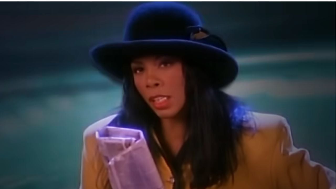 A woman with long dark hair wearing a black hat and yellow blazer sings into a vintage-style microphone against a blurred, dark background.