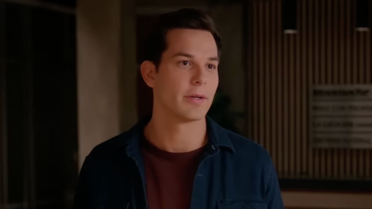 A man with short dark hair, wearing a dark blue shirt over a maroon t-shirt, stands indoors with a neutral expression. The background is softly lit and slightly blurred.
