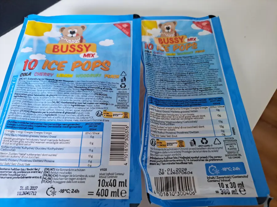 Two blue packs labeled “Bussy Mix 10 Ice Pops” with a bear graphic, showing flavors and nutritional info. The packs are placed on a white and black surface, one facing front and one showing the back details.