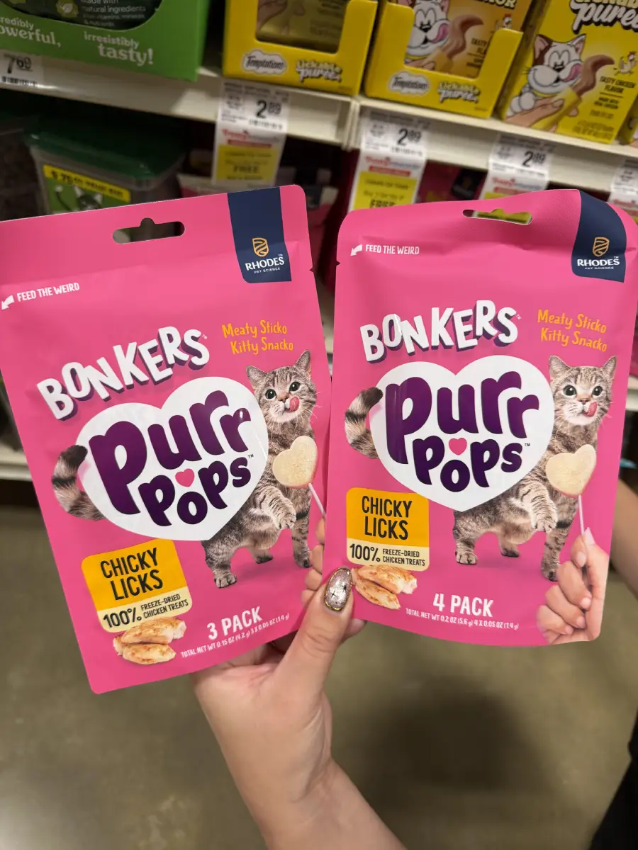 Two hands hold pink bags of Bonkers Purr Pops cat treats, featuring a cat image. One is a 3-pack, the other a 4-pack. Store shelves with other pet treats are visible in the background.
