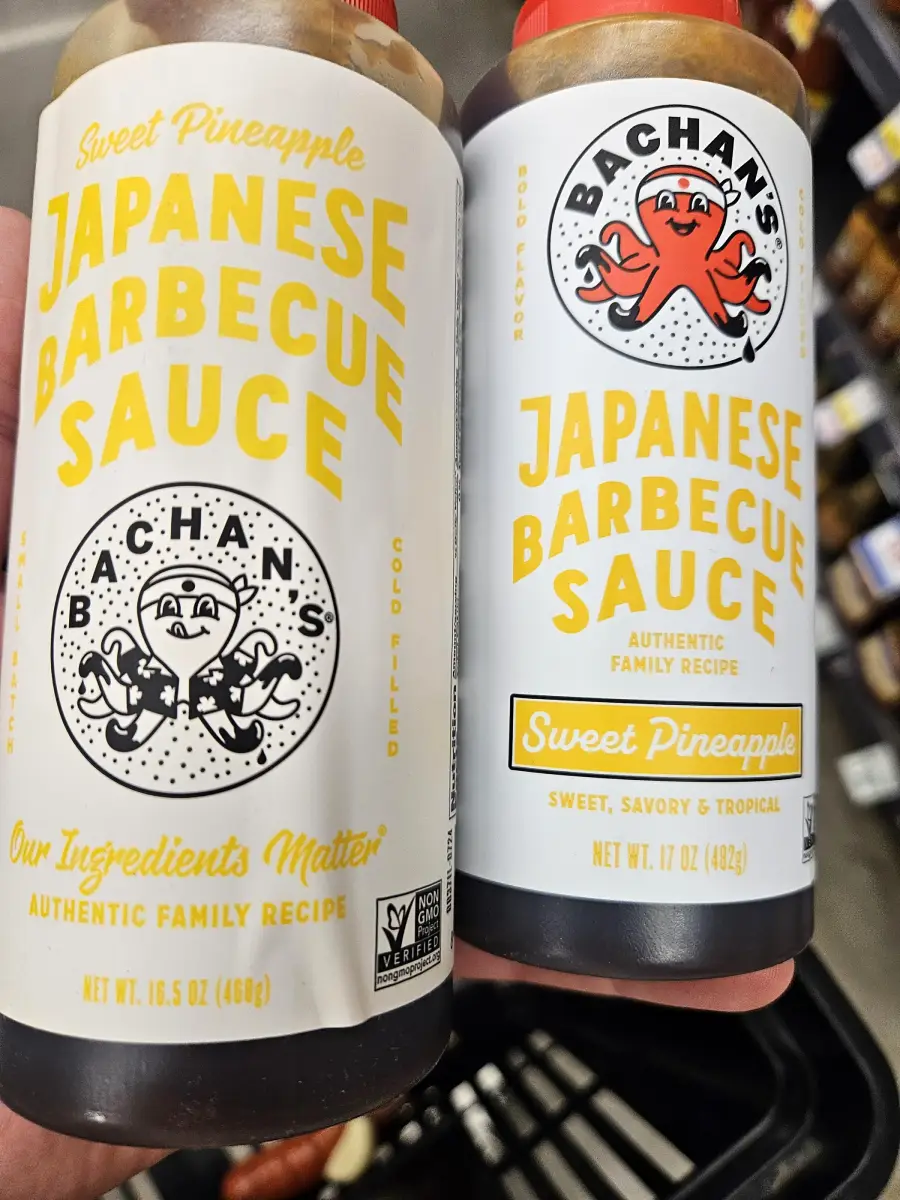 Two bottles of Bachan’s Japanese Barbecue Sauce, Sweet Pineapple flavor, are held side by side in a grocery store. The labels are yellow and white with an octopus logo and product information visible.