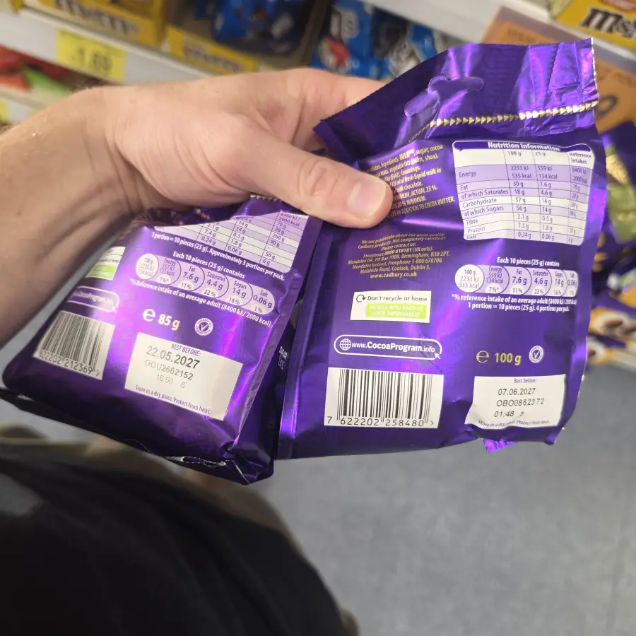 A person holds two purple chocolate packages in a store, comparing ingredients and nutritional info on the backs. Shelves with other products are visible in the background.