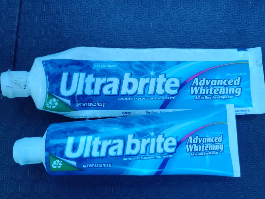 Two tubes of Ultra Brite Advanced Whitening toothpaste, one larger and one smaller, are placed on a dark textured surface. Both tubes are blue and white with green accents and white caps.
