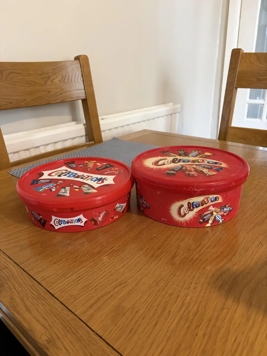 Two red Celebrations chocolate tubs are on a wooden table with two wooden chairs in the background.