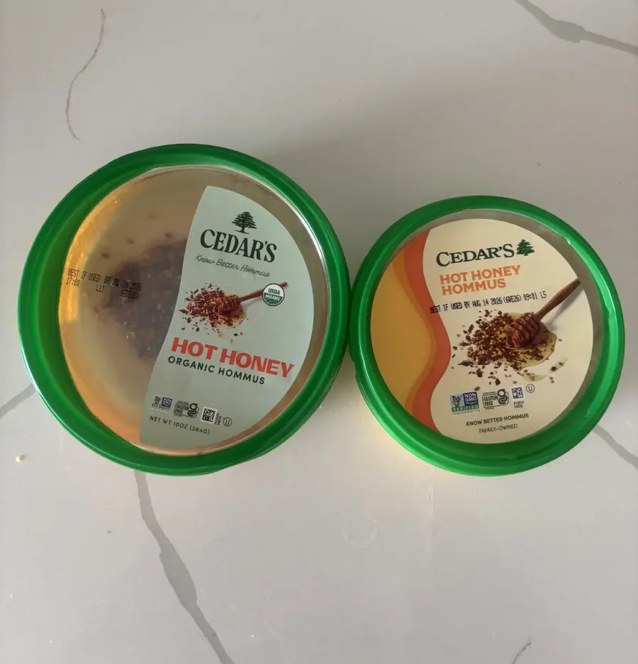Two Cedar’s brand containers of hot honey hummus with green lids are placed on a white surface. The left container shows the hummus inside, while the right container’s lid is fully labeled.