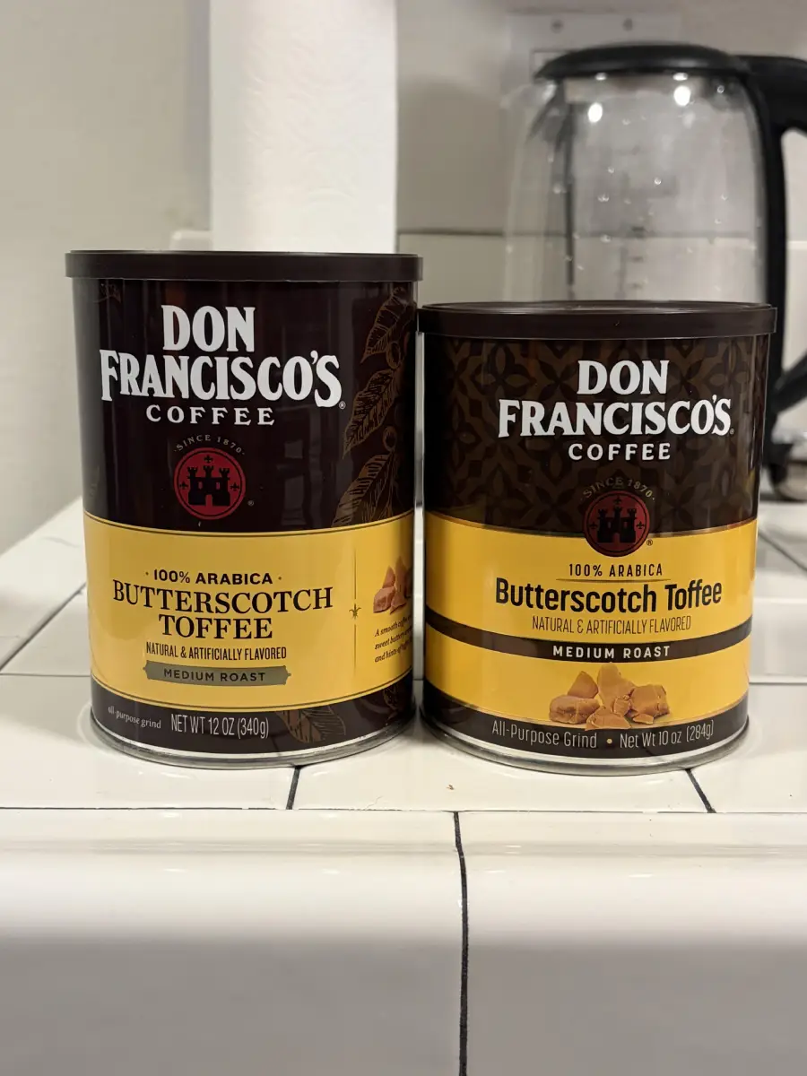Two cans of Don Francisco’s Coffee Butterscotch Toffee flavor sit on a tiled kitchen counter. The larger can is 12 oz, the smaller is 10 oz. A coffee maker is visible in the background.