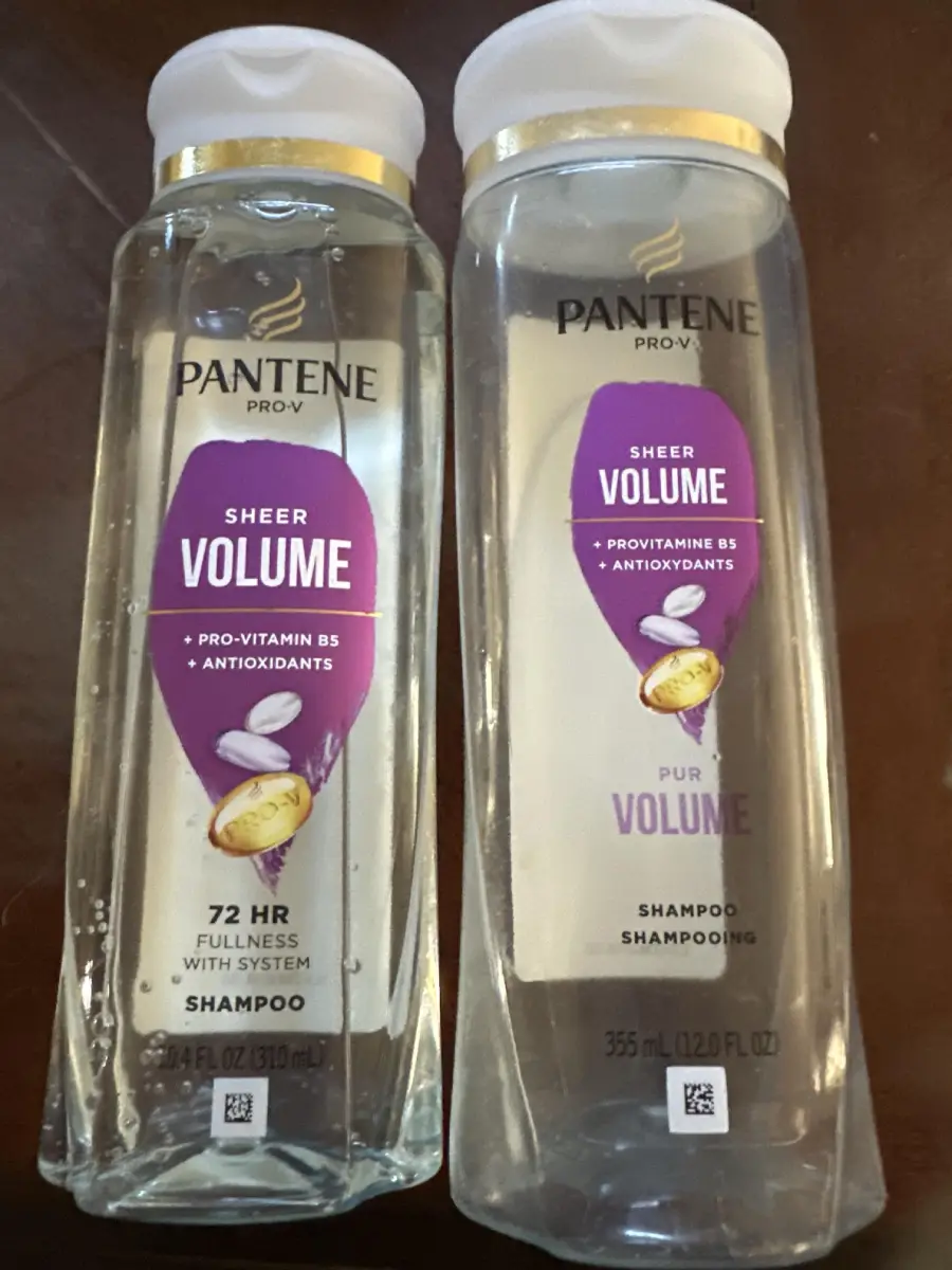 Two bottles of Pantene Pro-V Sheer Volume shampoo are shown side by side on a table. Both have gold and white caps and purple labels that mention pro-vitamin B5, antioxidants, and volume benefits.