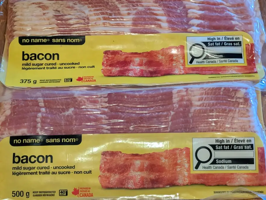 Two packages of No Name brand bacon, labeled “mild sugar cured: uncooked,” are shown. Each package highlights that the bacon is high in salt and fat, with Health Canada warnings on the front.