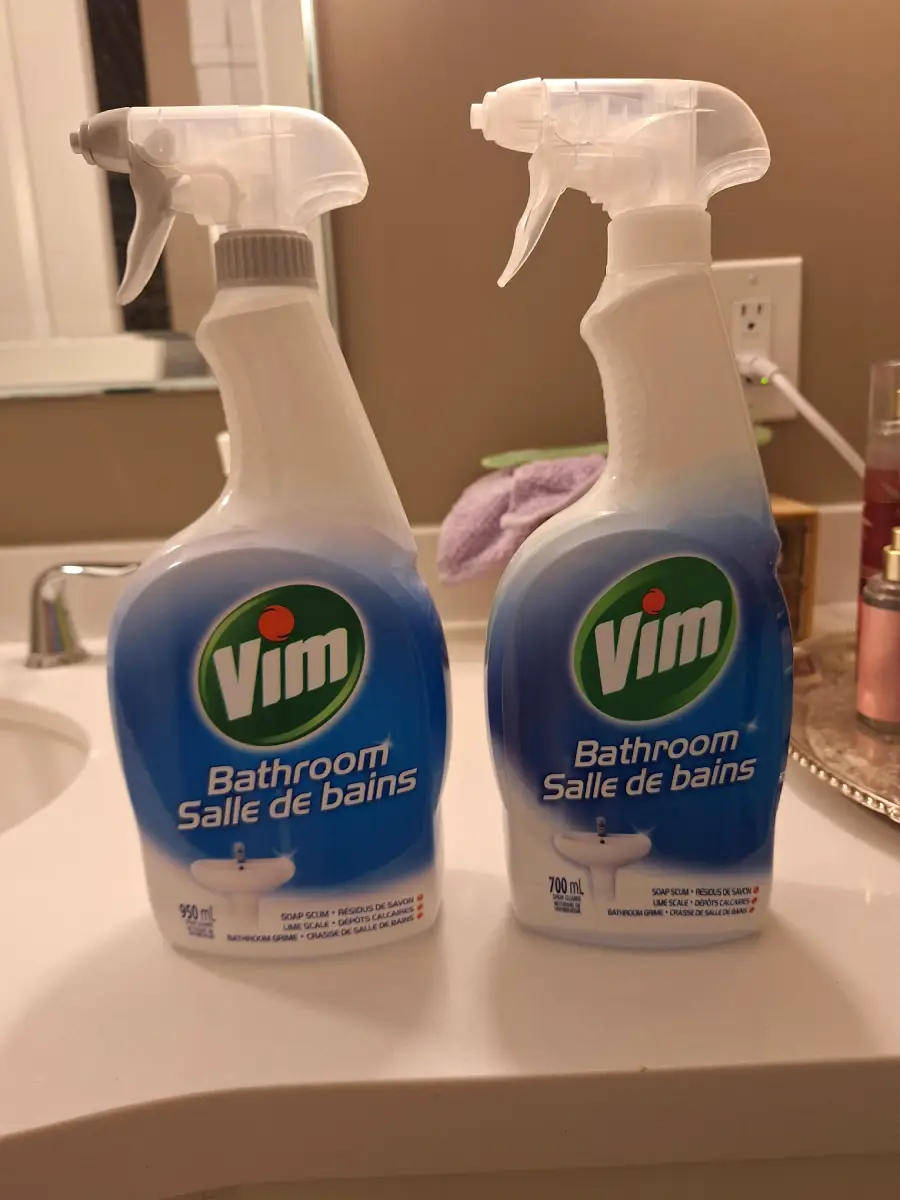 Two bottles of Vim Bathroom cleaner are placed side by side on a bathroom counter near a sink, a mirror, and a tray with various toiletries in the background.