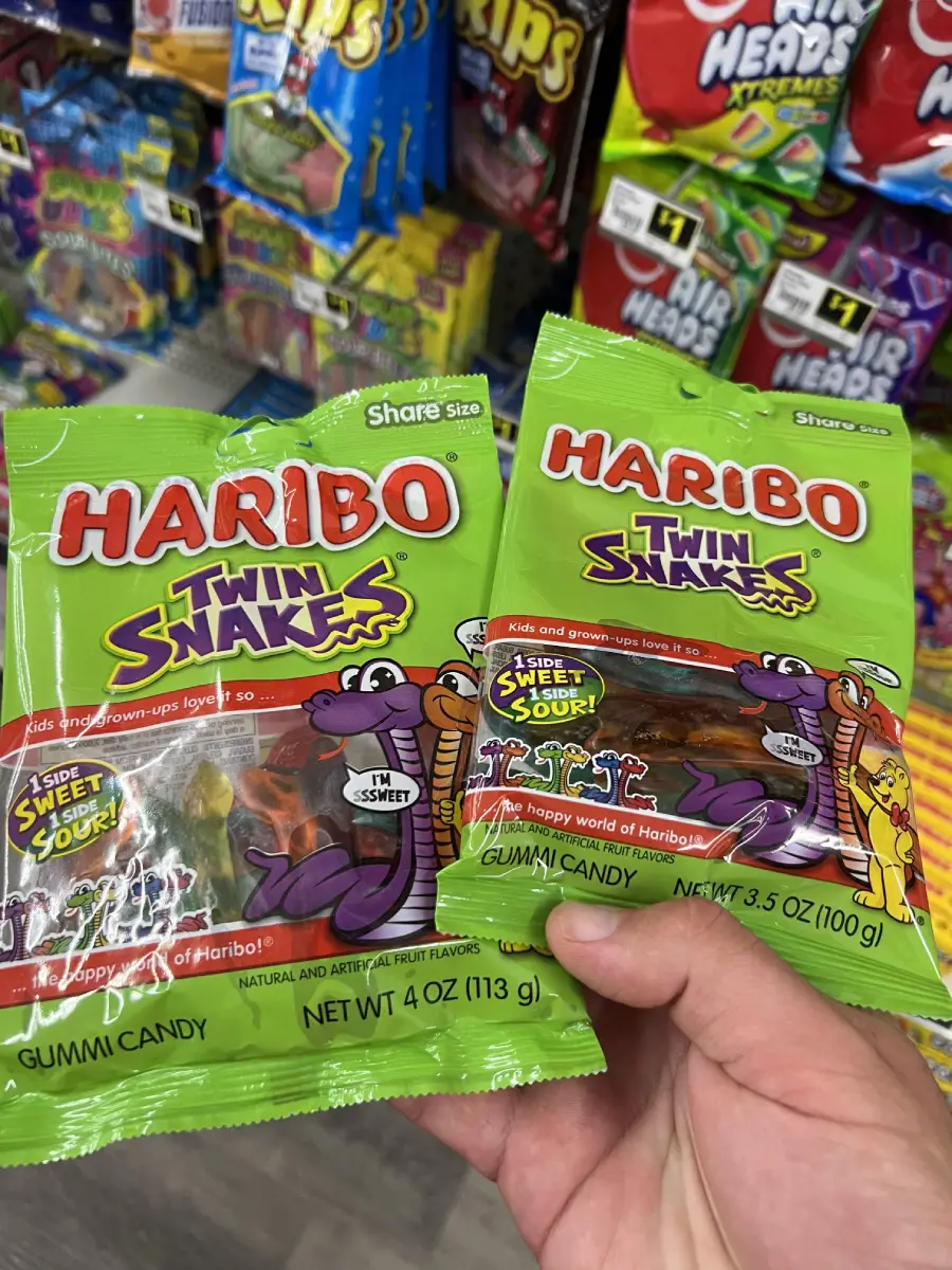 A hand holds two bags of Haribo Twin Snakes gummi candy in a store aisle, with colorful candy packages displayed in the background on shelves.