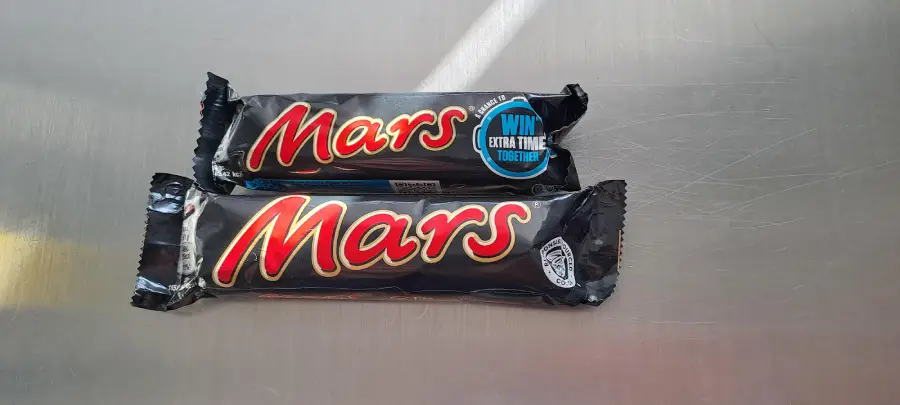 Two Mars chocolate bars in black wrappers are placed on a metallic surface. The upper bar has a blue label that says “WIN EXTRA TIME TOGETHER.” Light reflects on the right side of the image.