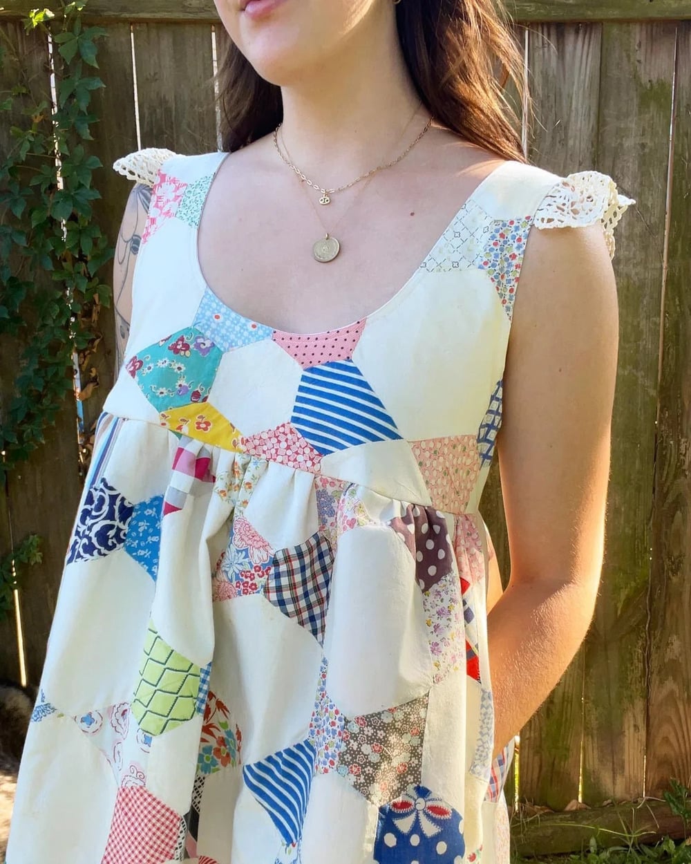 A woman wearing a sleeveless dress made from colorful patchwork fabric stands outside near a wooden fence with greenery.